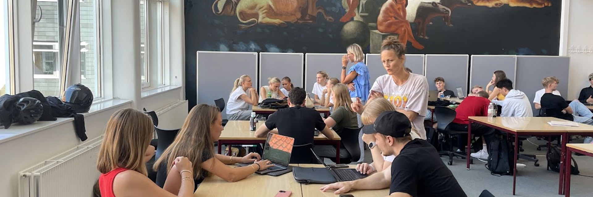 Grupper af elever sidder ved borde og arbejder med opgave, gruppe i forgrunden samtaler med facilitator